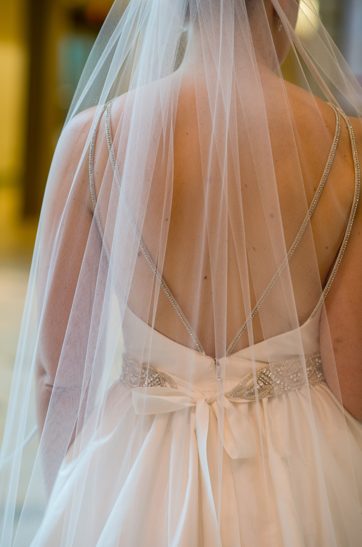 Close-up back detail of the bride in a low open back wedding gown with beaded straps, a belt and a tie with a veil overtop.