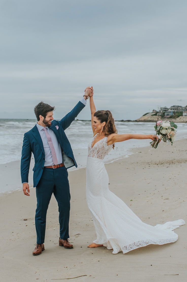 Bride in a fit and flare lace gown with a V-neckline, holding a bouquet and posing with groom in a blue suit, on a beach.