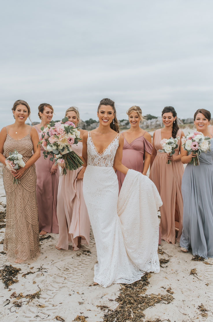 Bride smiles posing in a lace fitted gown with a V-neck, holding her train and a bouquet near bridesmaids on a sandy beach.