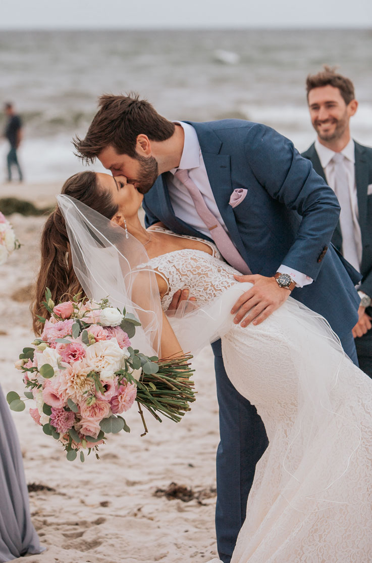 Groom dips his bride in a V-neck lace wedding gown with a veil, holding a pink bouquet as they kiss by the ocean.