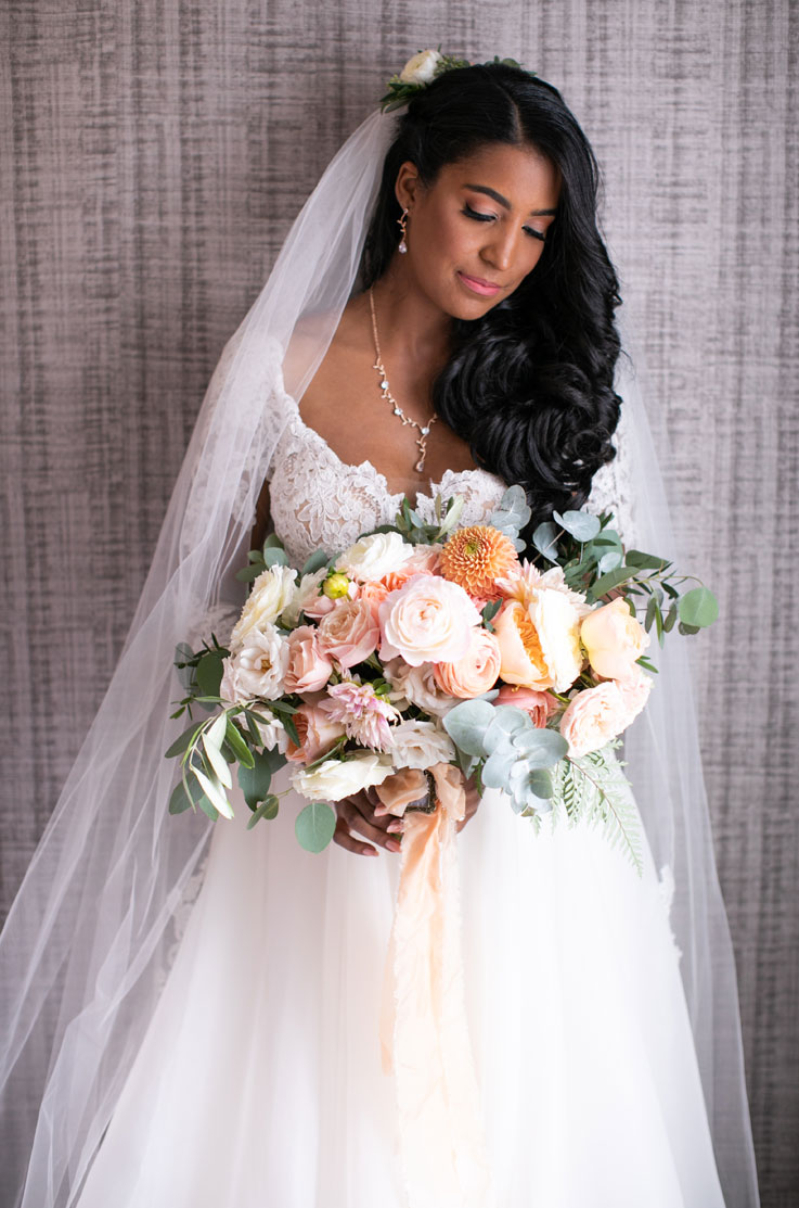 Close-up of bride in lace and organdy gown with a long veil and is holding a blush bouquet, posing by a textured wall.