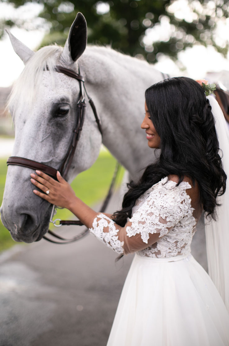 Bride in a tulle and lace bodice wedding dress with a full organdy skirt and sleeves, pets a white horse in a countryside.