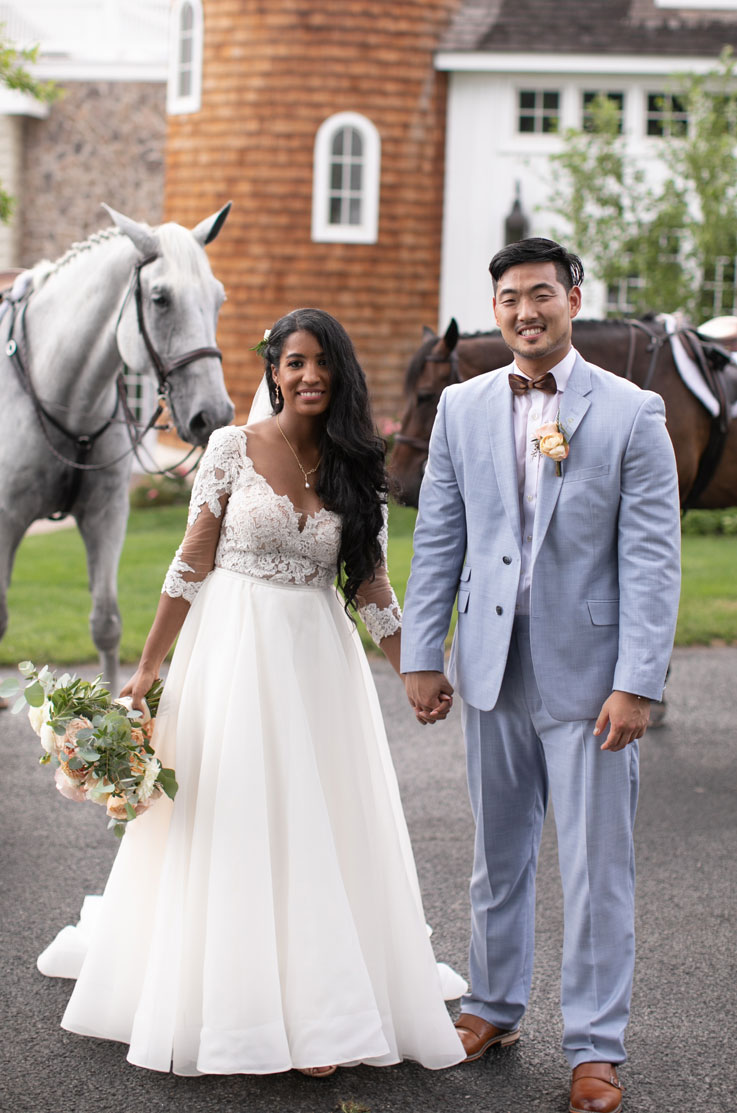 Bride in a lace and tulle gown with sheer sleeves and a full skirt, holds hands with groom in front of a venue with horses.