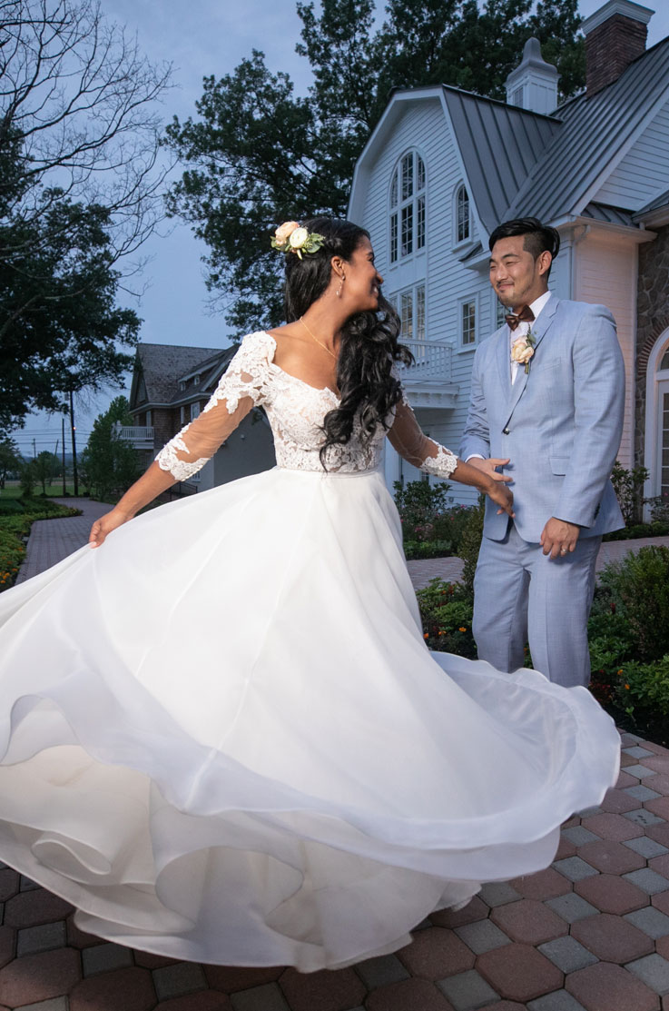 Bride twirls in an organdy and lace gown with tulle on the bodice and sleeves, facing groom in a blue suit outside the venue.