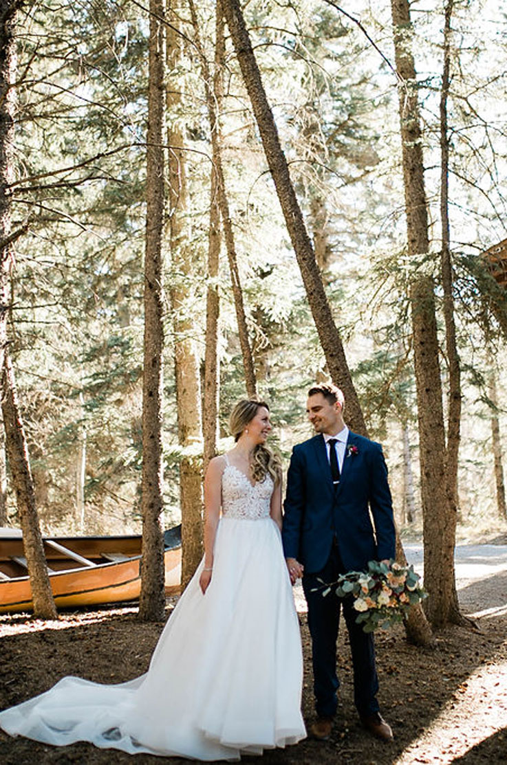 Bride in a lace bodice wedding dress with a full skirt, poses beside groom in a blue suit with flowers, in a forest setting.