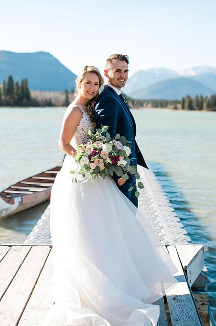 Bride in a sleeveless lace gown with a full organza skirt, on a lakeside dock and is holding a large bouquet beside groom.