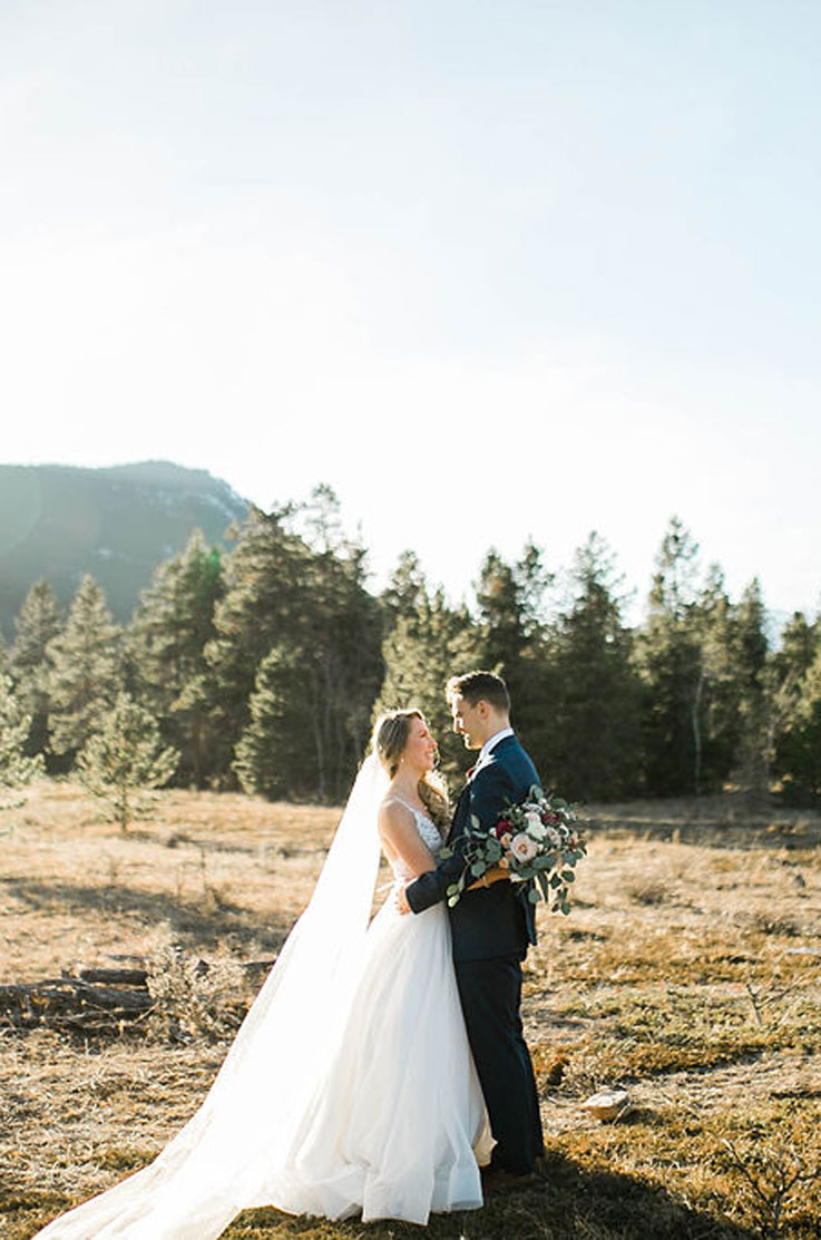 Bride in a lace and organza wedding gown with a long veil, embracing groom and holding a bouquet in front of evergreen trees.