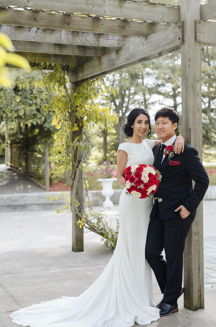 Bride in a fit and flare crepe gown with cap sleeves, holding a rose bouquet and standing next to the groom under a pergola.