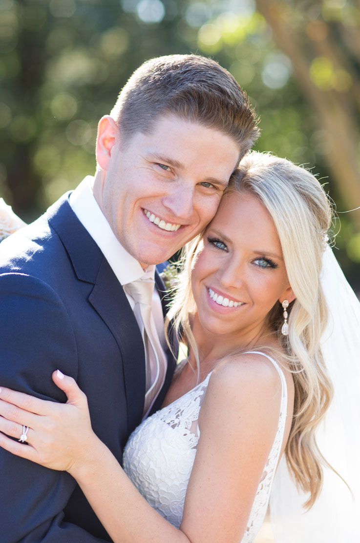 Bride in a sleeveless lace wedding gown and a veil, is smiling with her groom in navy suit and they hug outdoors.