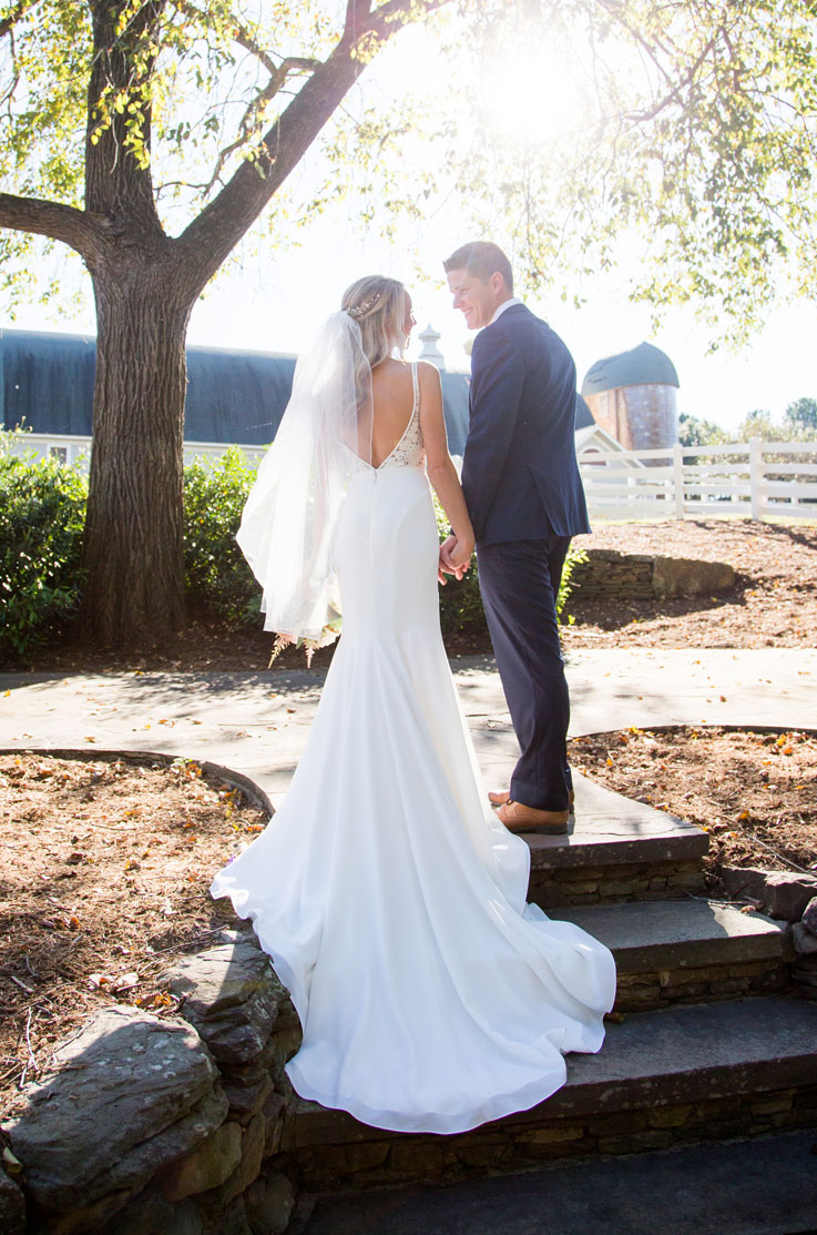 Bride in a deep-V back crepe gown with a veil and lace bodice, holding hands with groom in the sunlight on stone steps.