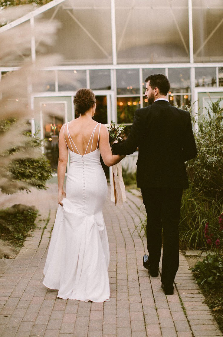 Bride in a sleeveless fit and flare gown with a low open back, walks next to groom on a garden path at a greenhouse.
