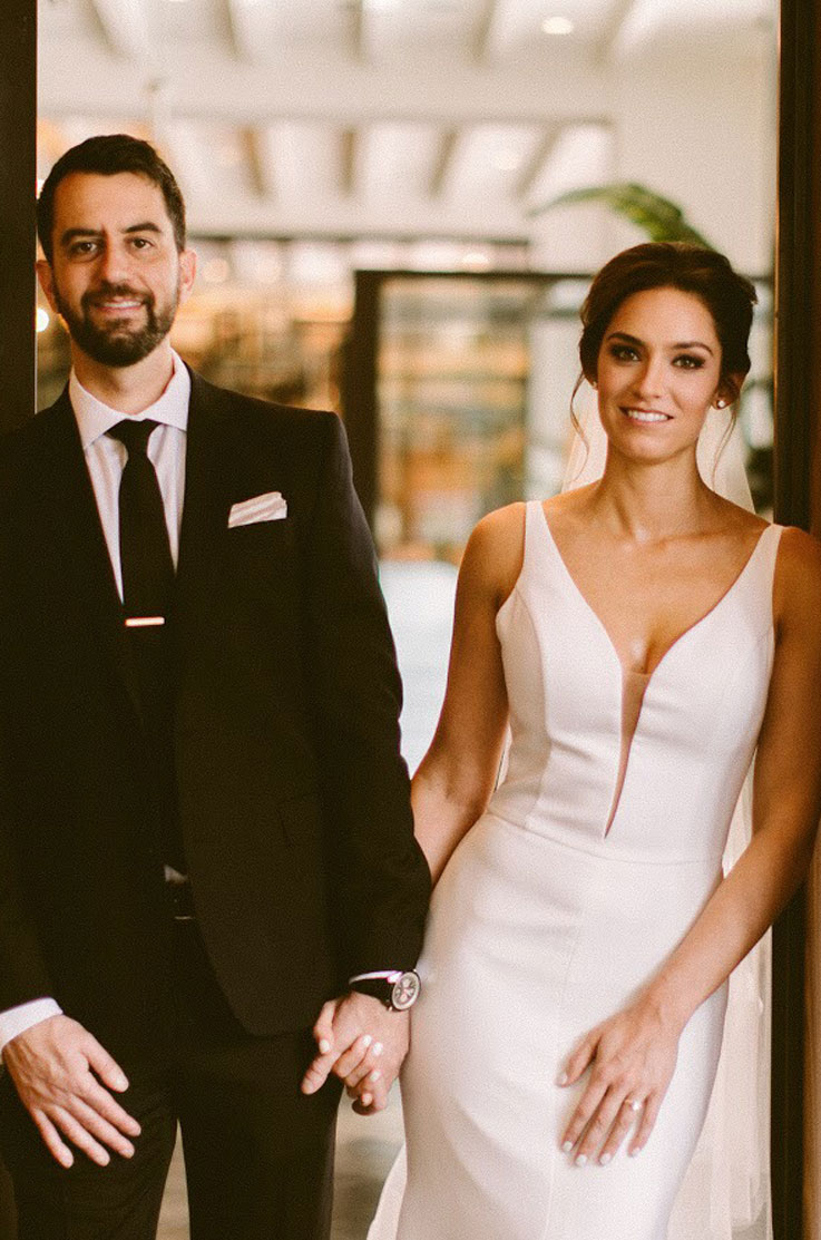 Bride in a crepe fit and flare gown with a plunging V-neckline, poses beside groom in a modern glass building.