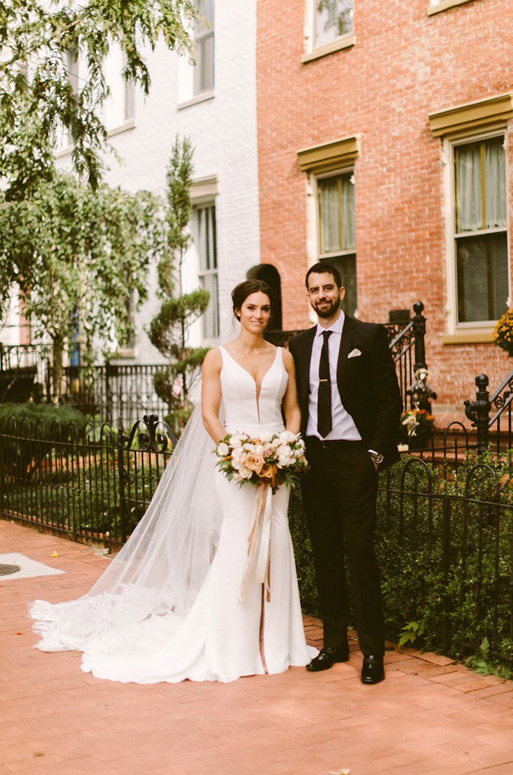 Bride stands beside groom, in front of buildings, in a crepe fit and flare gown with a long veil and is holding flowers.