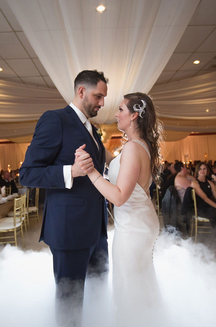 Groom dances with his bride wearing a satin fit and flare wedding dress, with a low back and beaded straps in low-lying fog.