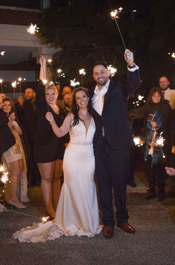 Bride in a satin plunging V-neck gown with a beaded belt, stands next to groom as they hold up sparklers at night with guests.