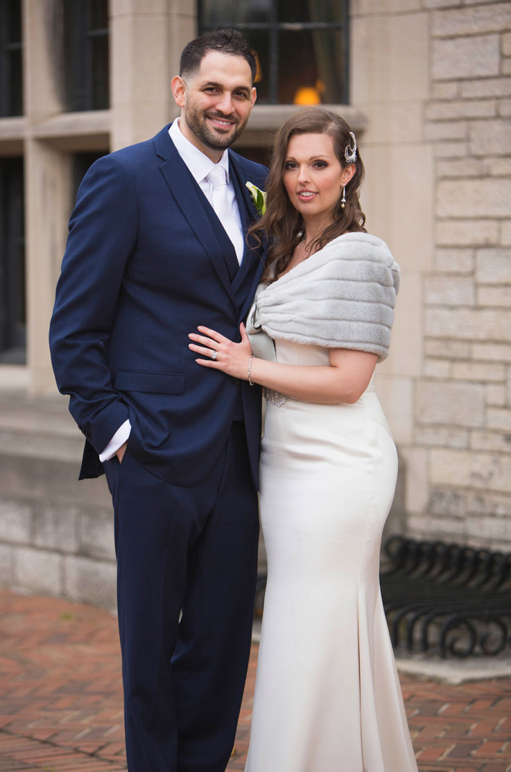 Bride poses in a fit and flare gown with a fur shawl overtop, beside groom in a blue suit in front of the venue.