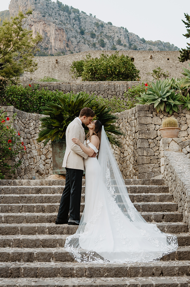 Groom kisses his bride in a fit and flare off-shoulder wedding dress with a lace-trim veil, on a stone staircase.