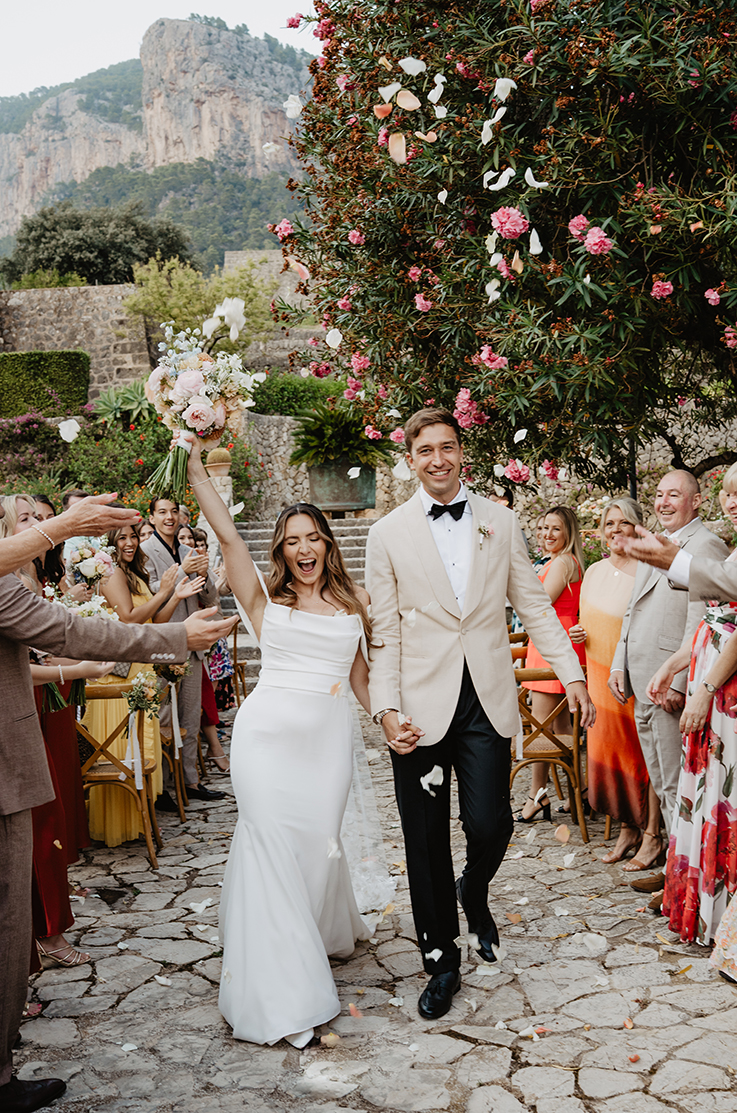 Bride in a fit and flare crepe gown, walks down the aisle with her groom, celebrating as guests throw flower petals in the air. 
