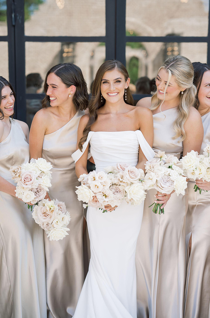 A bride in a wedding dress with draped sleeves is surrounded by bridesmaid in one-shoulder dresses, all smiling and holding bouquets, standing in front of a window.