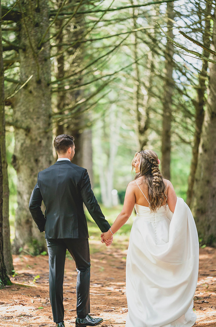 A bride in a draped bodice wedding dress and a groom in a black suit face away and walk hand in hand down a path under a canopy of trees.