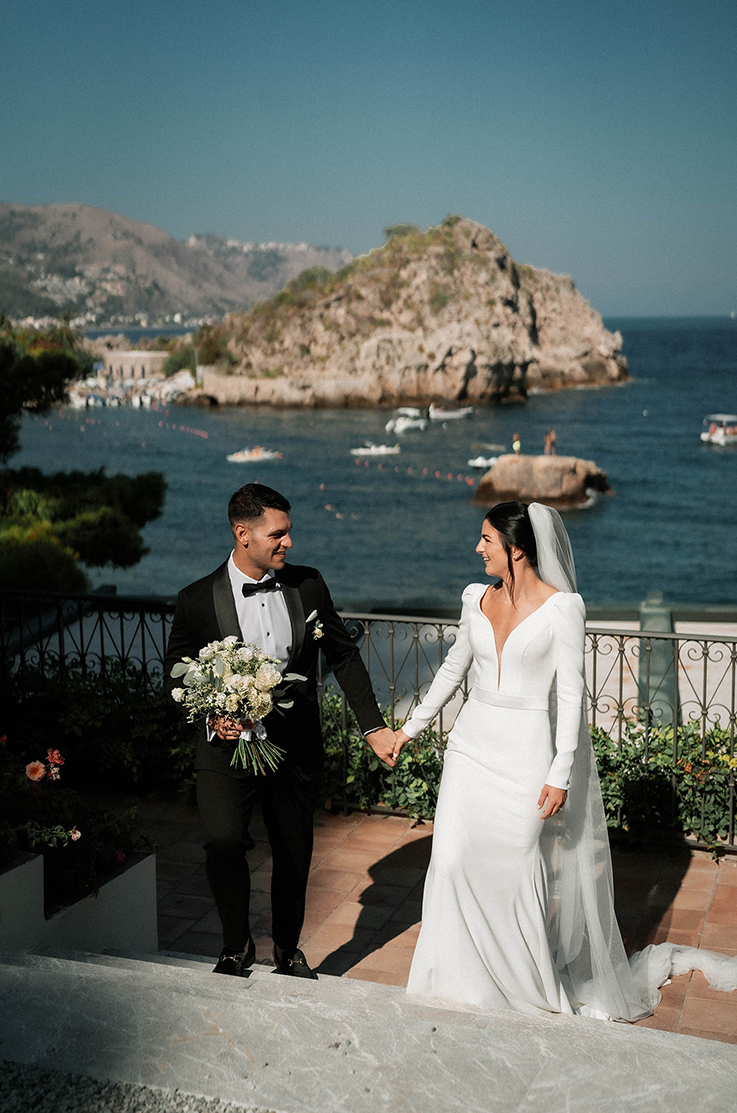 A bride in a long sleeve wedding dress and a groom in a black suit and bow tie, holding hands and climbing the stairs of a balcony overlooking a coastline and mountains.