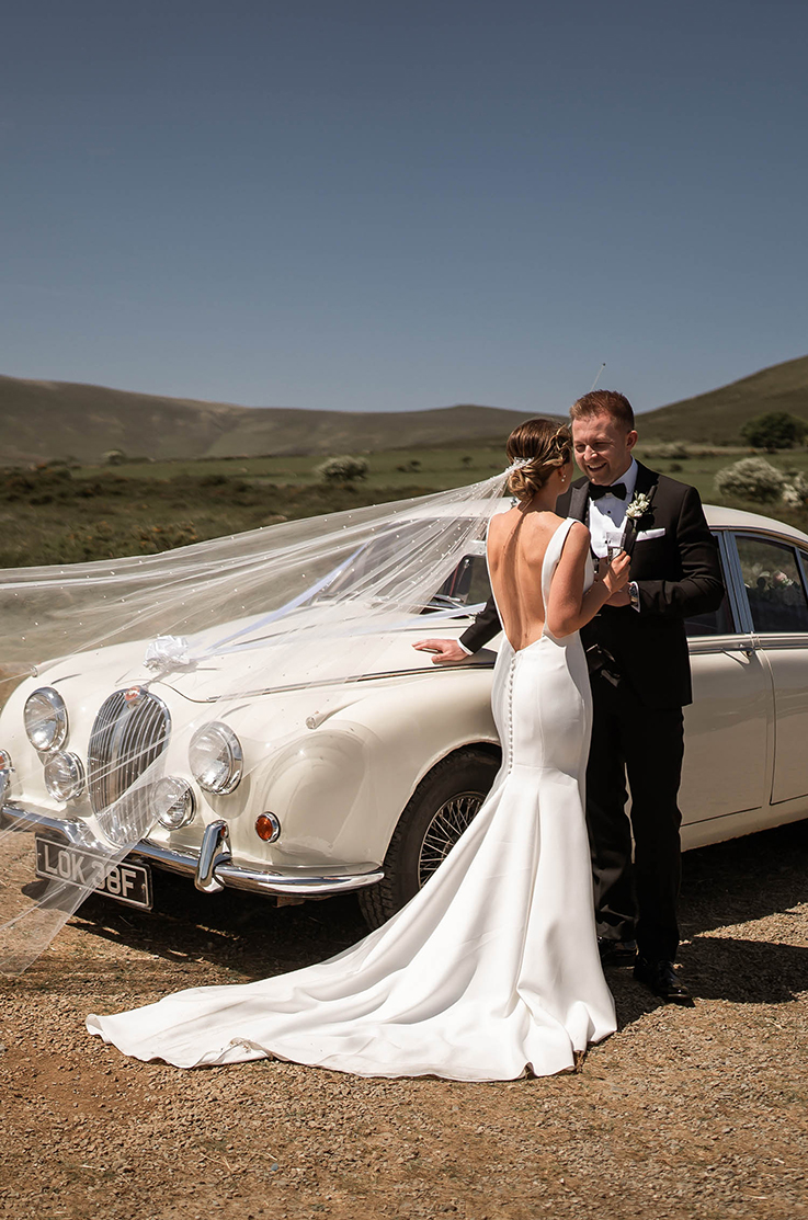 A bride in a sleeveless wedding dress wearing a veil faces a groom in a black suit and a bowtie, both of them leaning against a white classic car outside in front of rolling hills.
