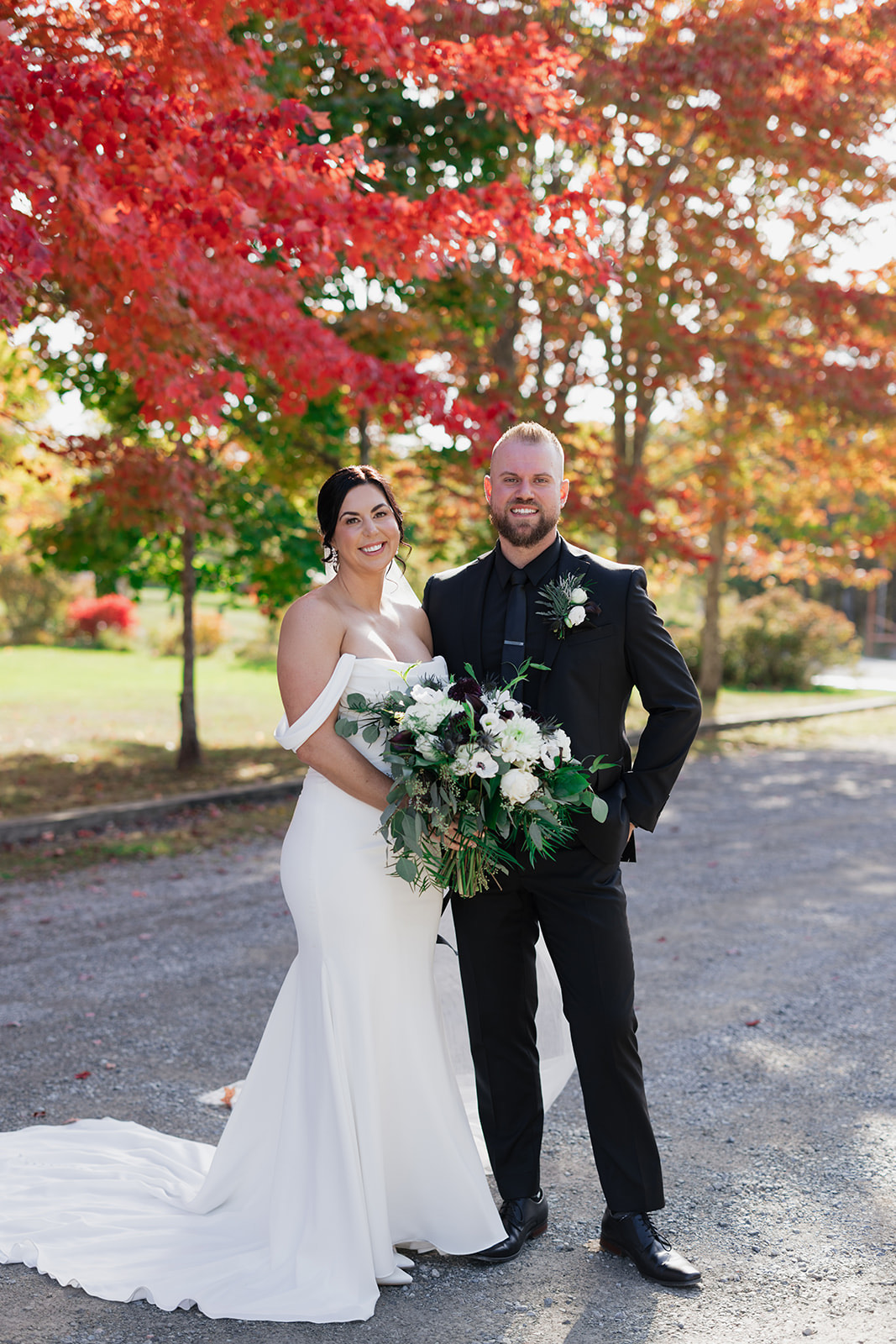 Bride in an off-shoulder fit and flare gown in crepe, is holding a bouquet next to the groom in a black suit outdoors.