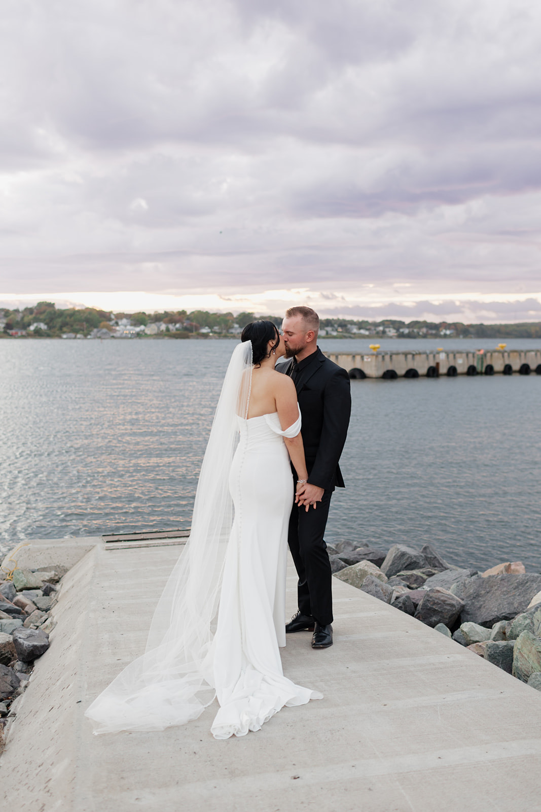 Groom kisses his bride in an off-the-shoulder crepe gown with a fit and flare skirt and veil, at a waterfront as the sun sets.