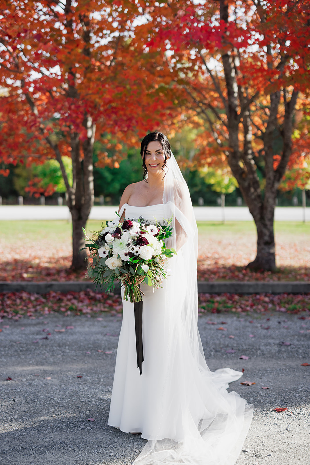 Smiling bride poses in front of red maple trees in a draped crepe bodice gown with a long veil and is holding a large bouquet.