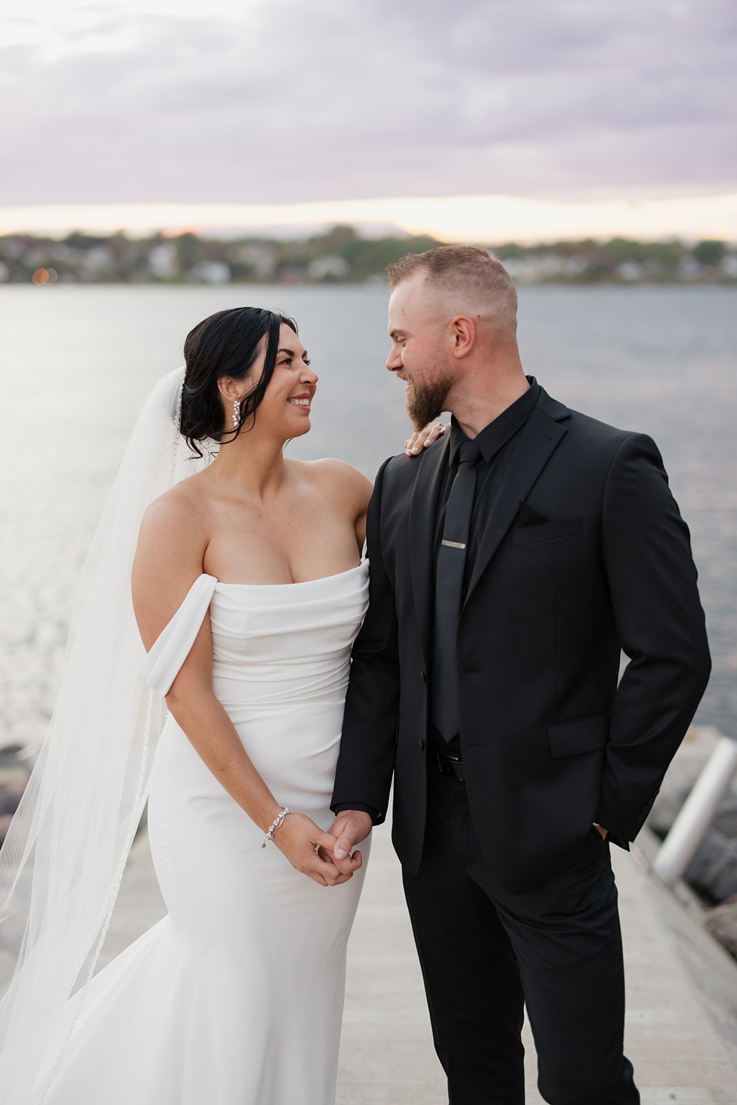 Bride smiles at the groom, wearing a crepe fit and flare wedding gown with off-shoulder sleeves and a veil, by a lakeside.