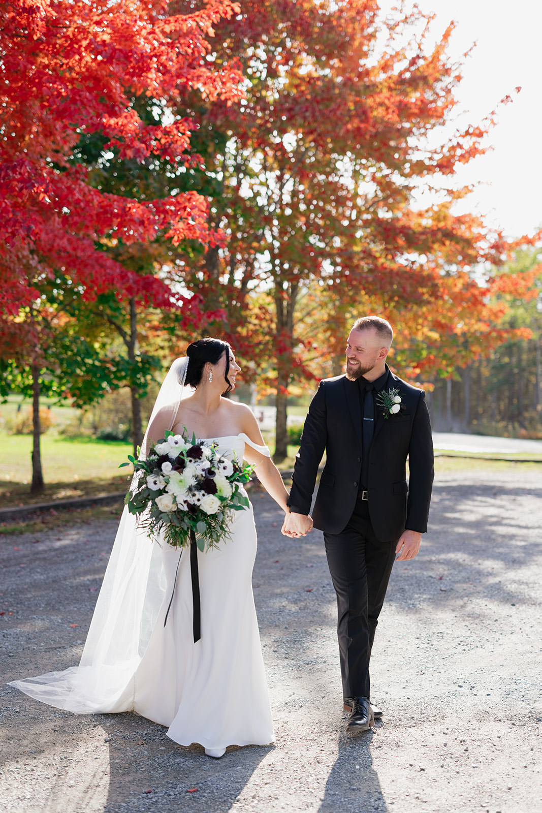 Bride in a crepe wedding dress with a veil and draped sleeves, holds a bouquet and walks alongside groom beneath fall trees.