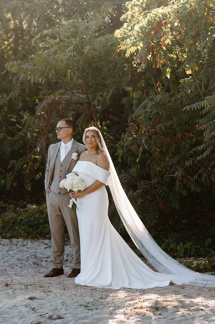 Bride in a crepe wedding dress with a portrait collar and a veil, is holding a rose bouquet next to the groom on a sandy beach.
