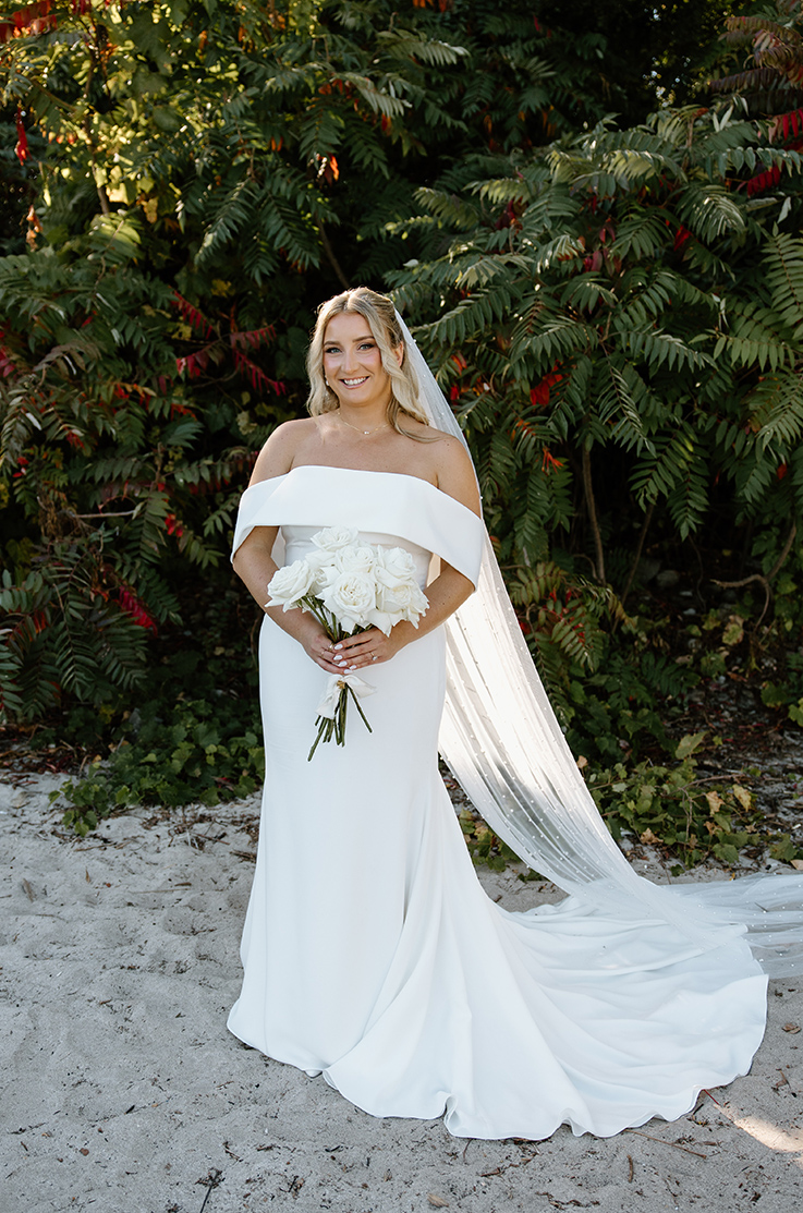  Bride poses in an off shoulder fit and flare gown with a veil, holding a white rose bouquet on a sand beach with greenery.