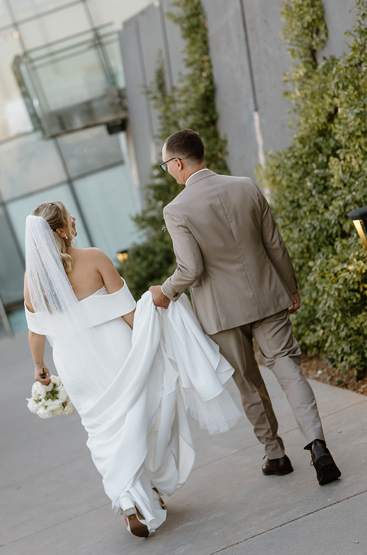 Bride in an off-shoulder crepe gown with a veil, is walking alongside her groom while he holds her train up outside the venue.