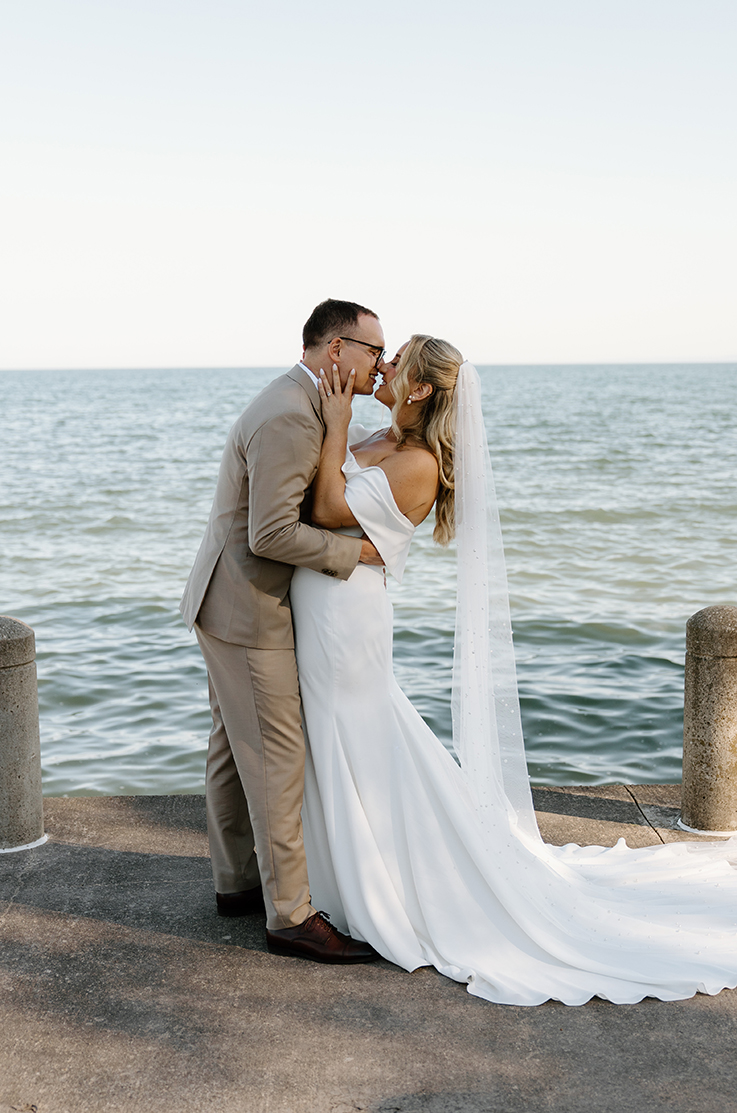 Groom embraces his bride in a crepe off-shoulder wedding gown with a veil and fit and flare skirt, in front of a lake.
