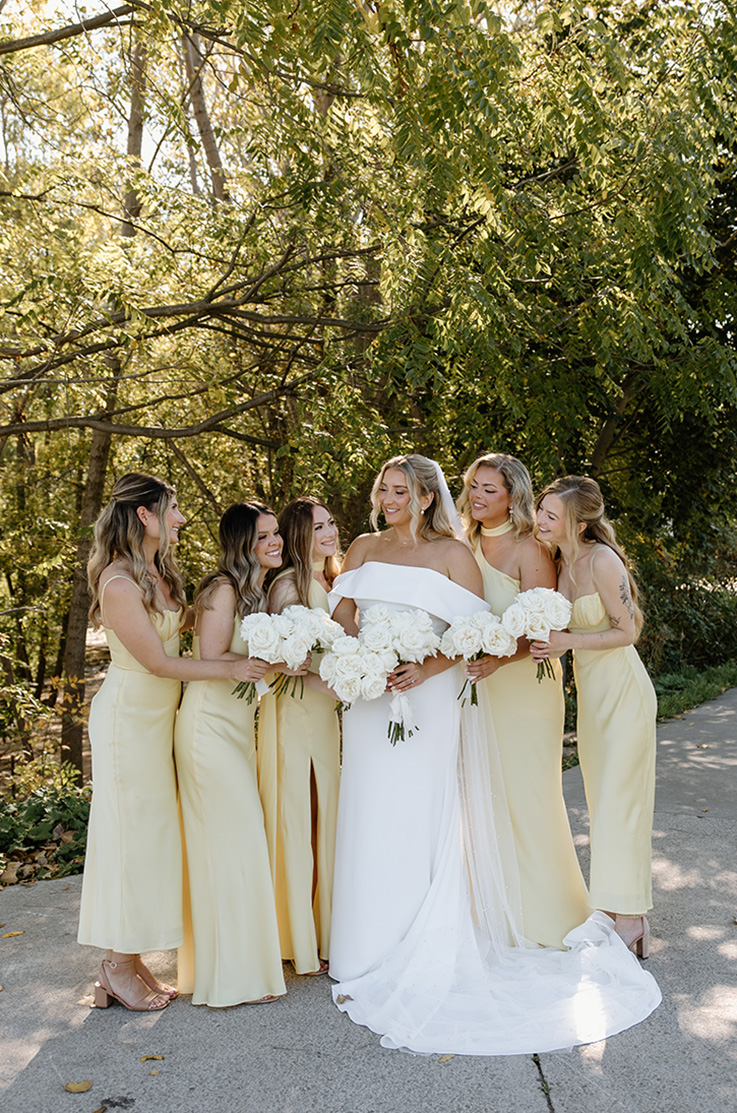 Bride in a crepe fit and flare gown with a portrait collar, smiles next to bridesmaids in yellow dresses with bouquets outdoors.