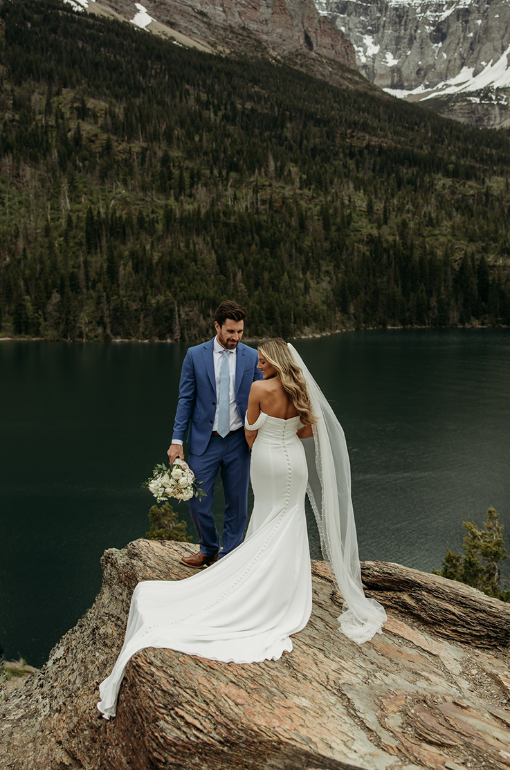 Bride poses on a large rock in a fitted draped sleeve wedding dress with a long veil, next to groom in front of mountains.