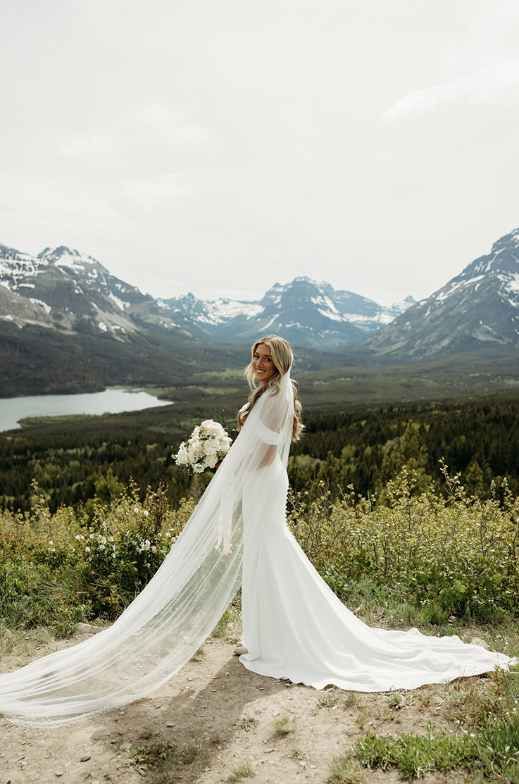 Bride smiles in a crepe draped bodice gown with off-the-shoulder sleeves and a long veil, posing on a mountain summit.
