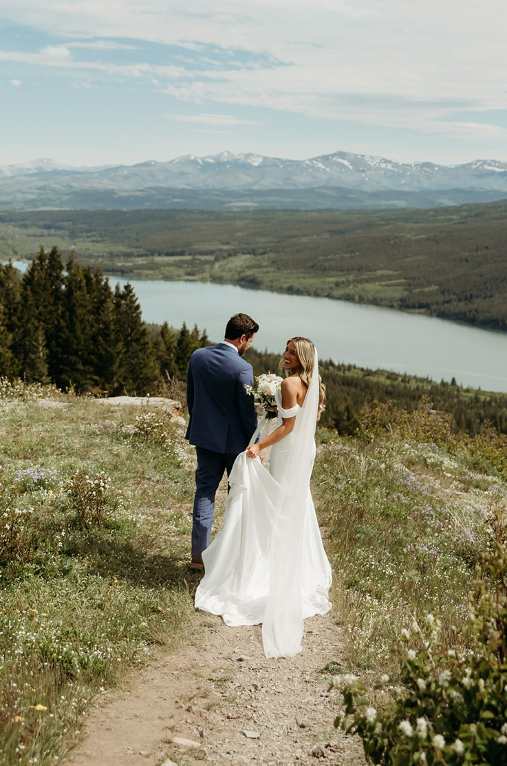 Bride in an off shoulder crepe gown with draped sleeves and a veil, walks beside groom holding her train, on a mountain trail.