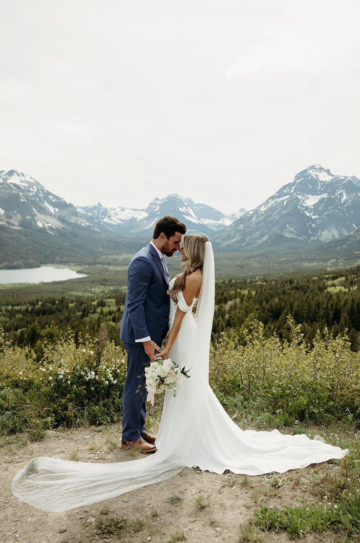 Bride in a draped off-shoulder wedding gown holds a white bouquet, and is facing groom in a blue suit on a mountaintop.