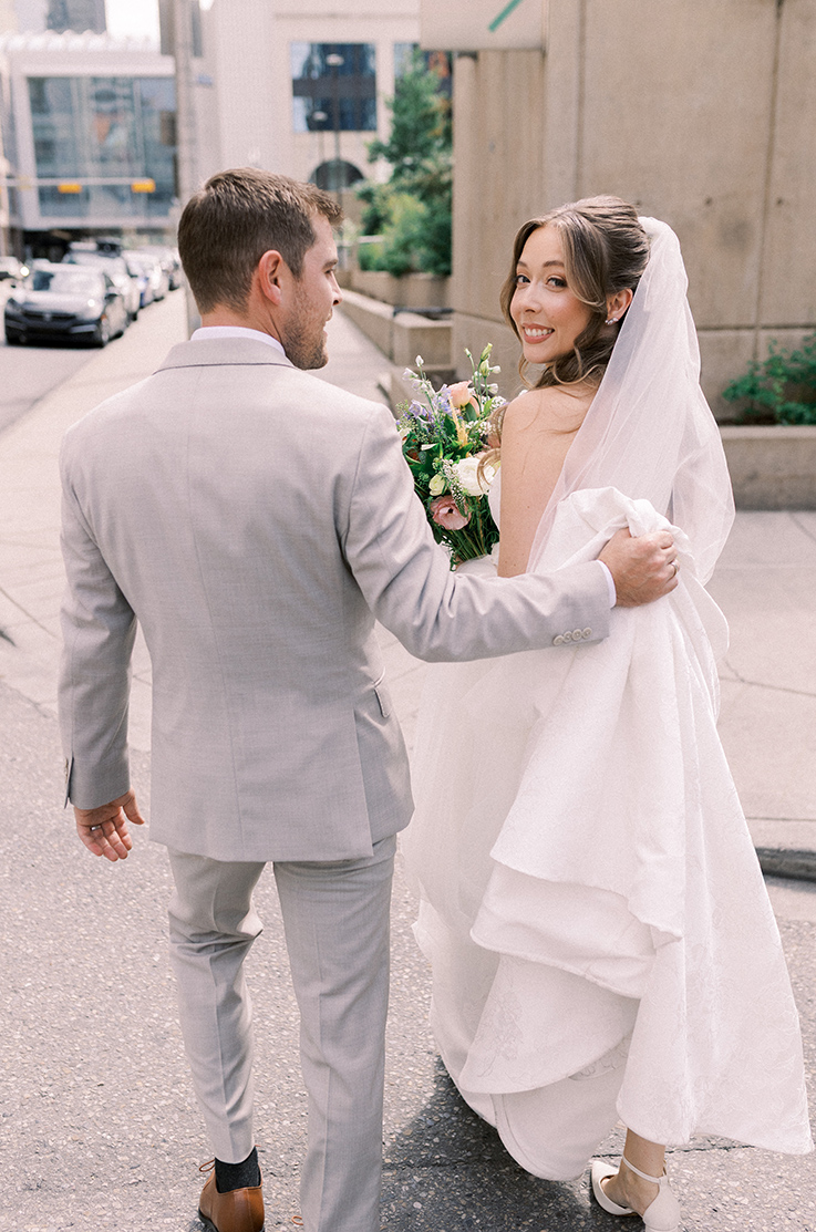 Bride looks over her shoulder in a Jacquard gown with a veil, smiling as groom picks up her train and they walk down a street.