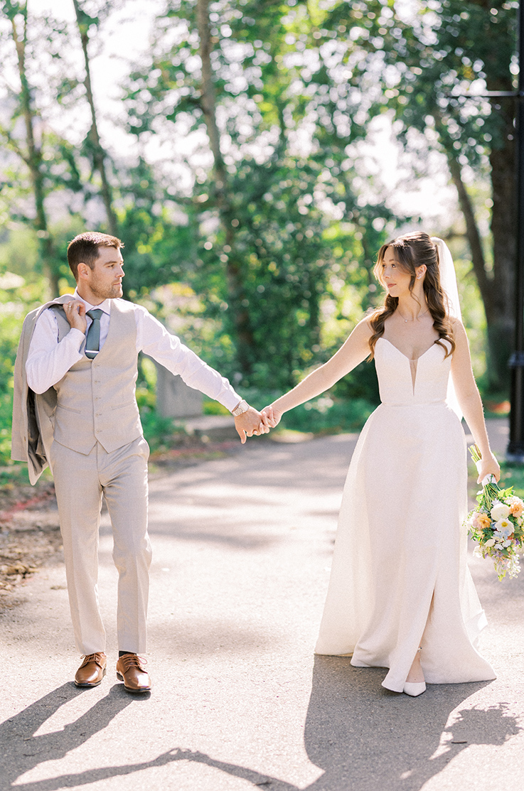 Bride in a strapless A-line gown with a veil, holds hands with her groom in a tan suit, on a sunlight tree-lined path.