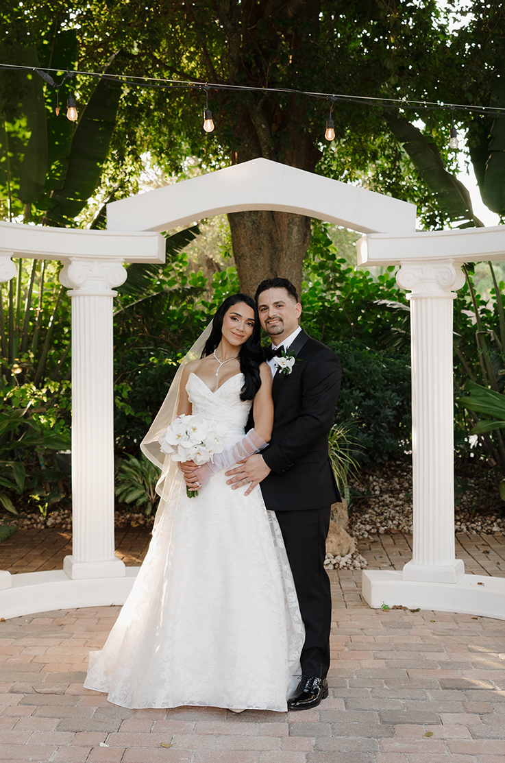 Groom poses with bride in a strapless burnout organza wedding dress with a sweetheart neck, at the altar beneath string lights.