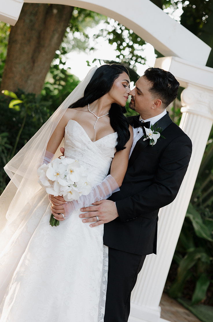 Groom embraces his bride in a sweetheart neck organza gown with sleeves and a veil, under a white stone archway.