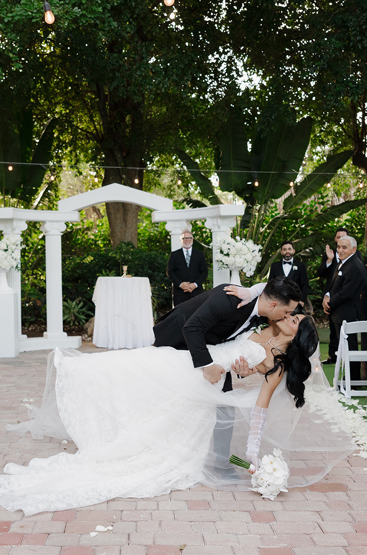 Groom dips his bride in a strapless burnout organza gown with sheer sleeves and a veil holding a white bouquet outdoors.