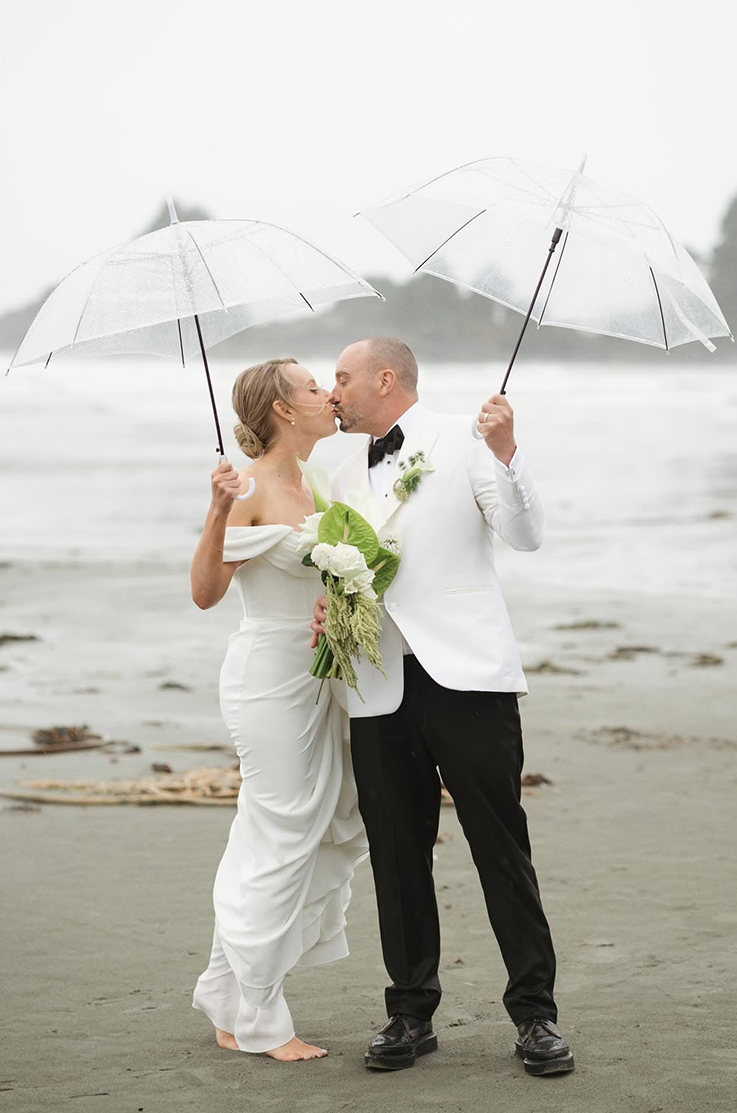 Bride in a draped fit and flare wedding dress, with off-shoulder sleeves, kisses groom under clear umbrellas on a rainy beach.