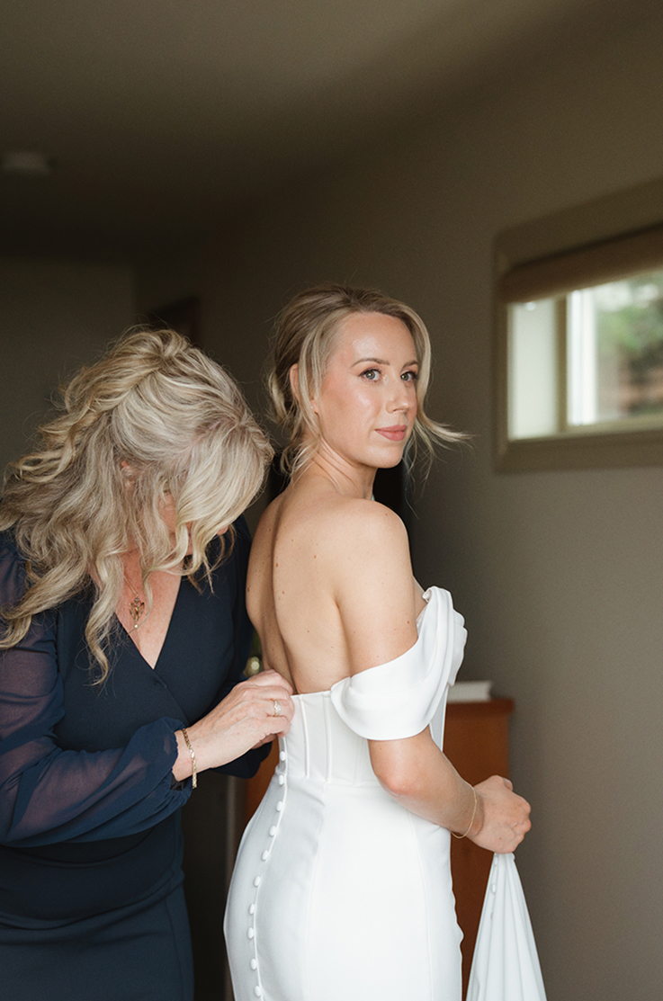 Bride is fastened into an off-shoulder fit and flare crepe gown with a corset bodice and buttons, indoors by a bright window.