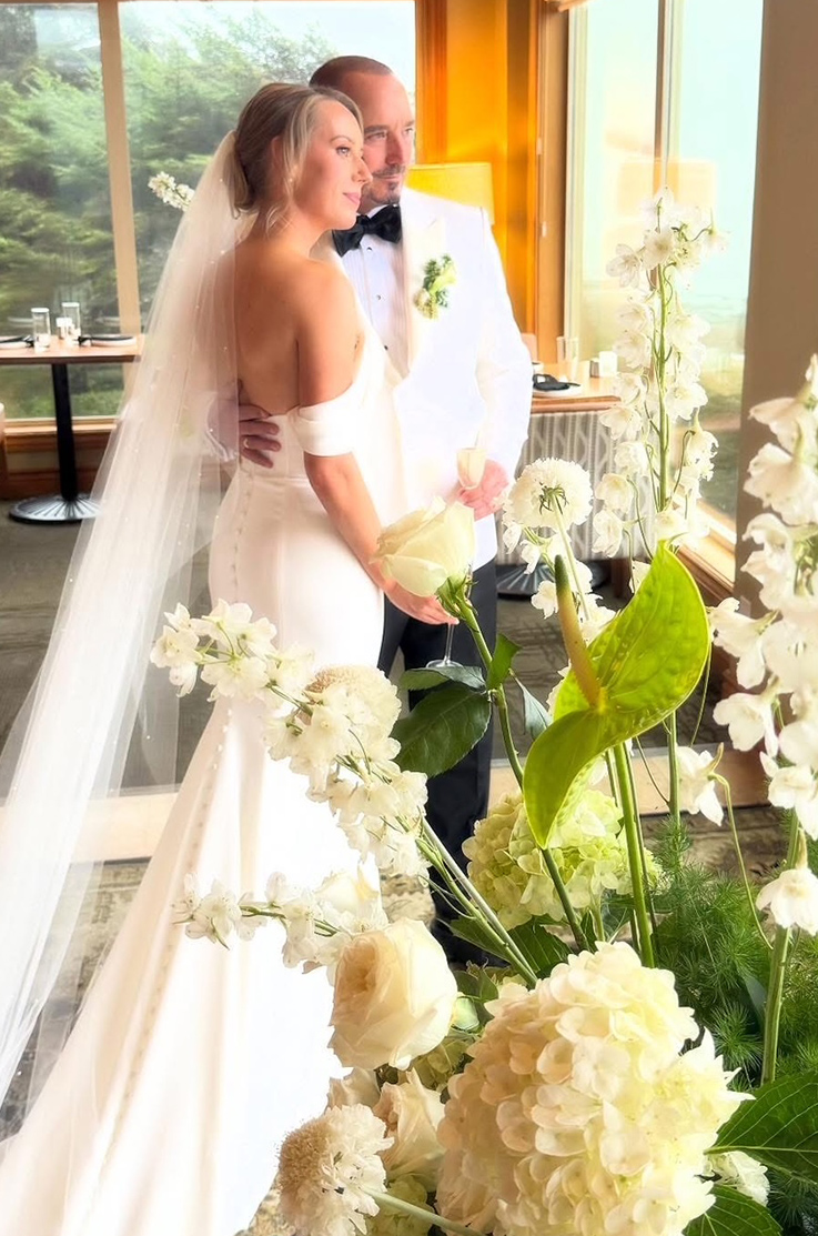 Bride smiles in an off-shoulder crepe gown with a buttoned back, next to groom and white flowers, in front of large windows.