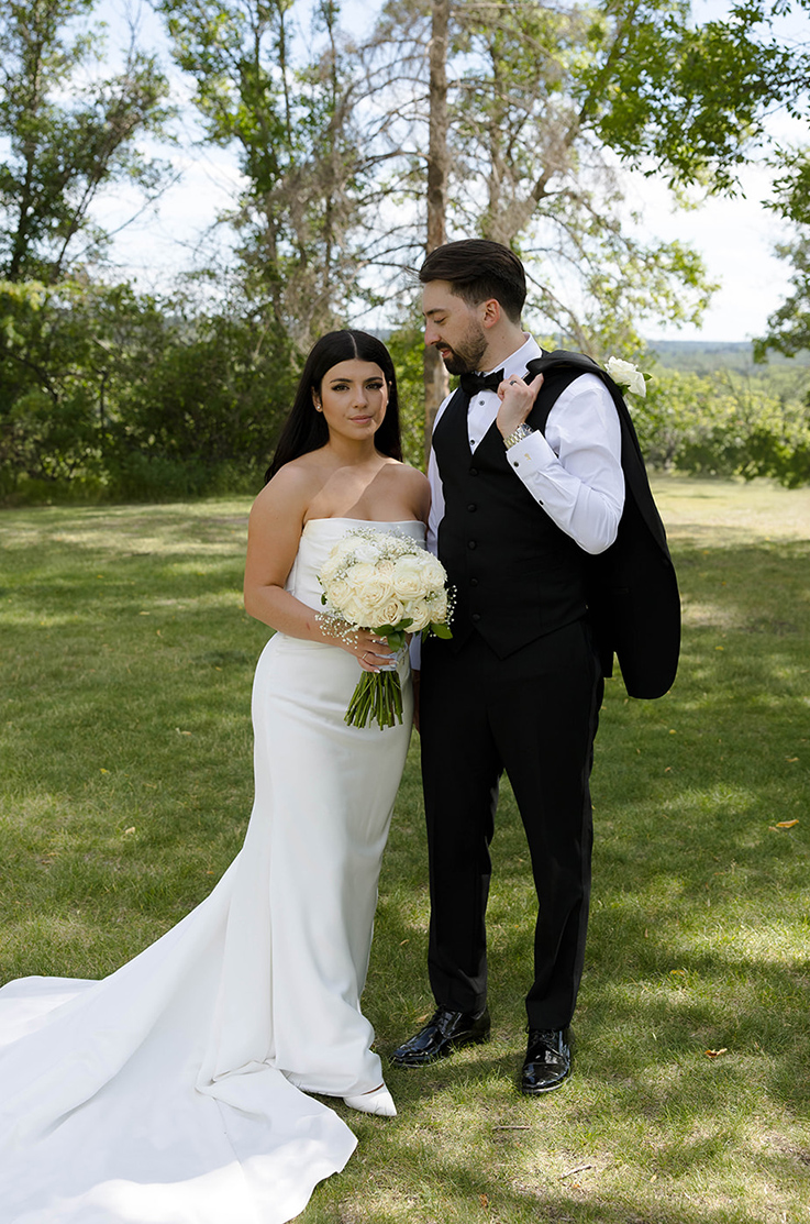 Bride in a strapless satin wedding dress poses and is holding a rose bouquet, next to her groom in a sunny outdoor area. 