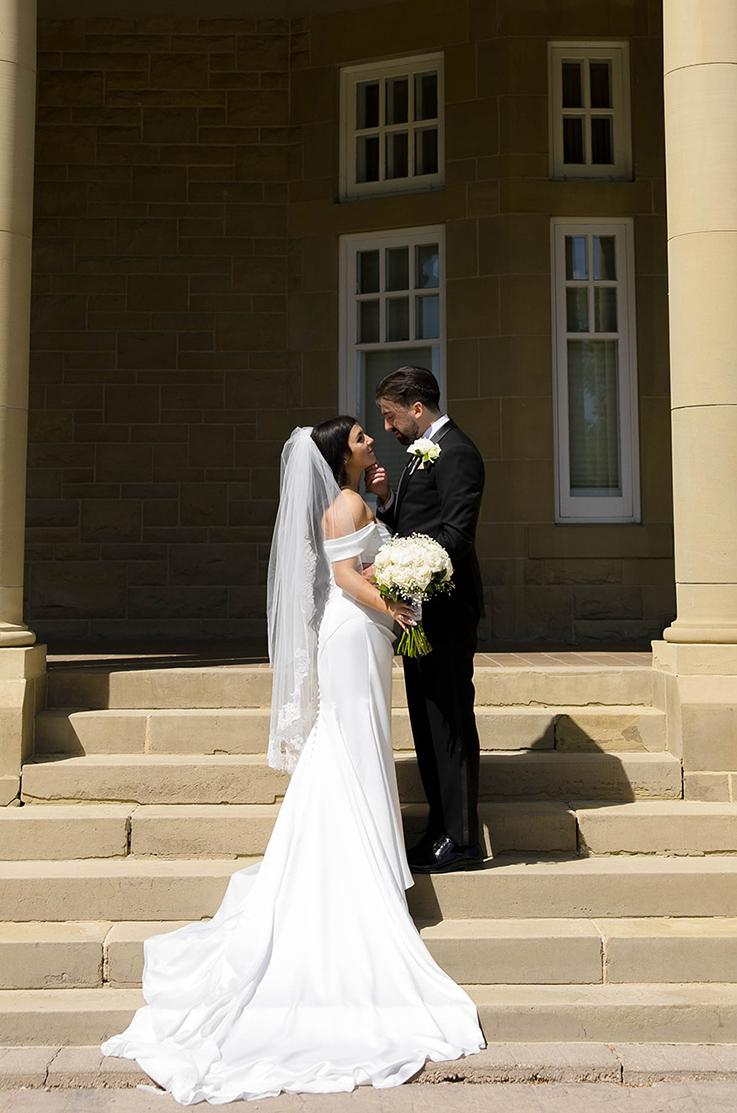 Bride in a satin fit and flare gown with a veil, holds a bouquet and faces her groom on stone steps next to tall pillars.